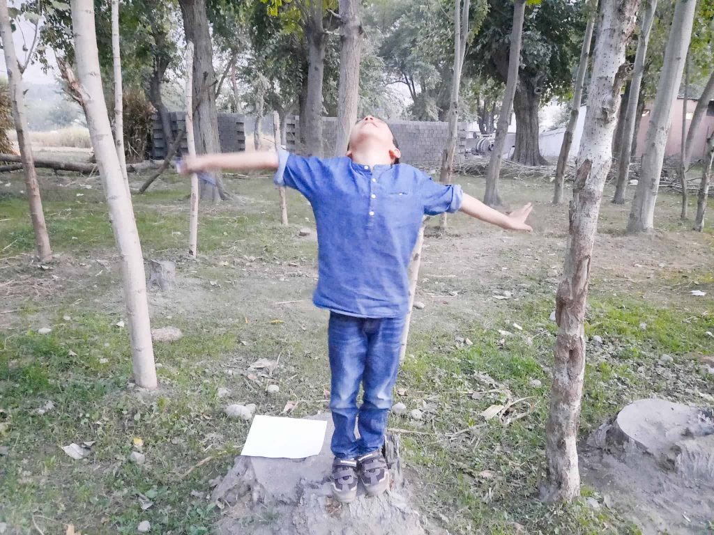 child standing on a tree stump with arms open to pretend he is the rest of the tree, while the fall scavenger hunt printable lies on the stump by the child's feet